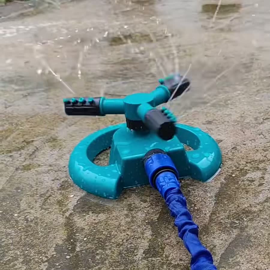 Aspersor giratorio de tres brazos en marcha sobre el suelo, con la manguera conectada a la base: los brazos giran y el agua sale por sus puntas