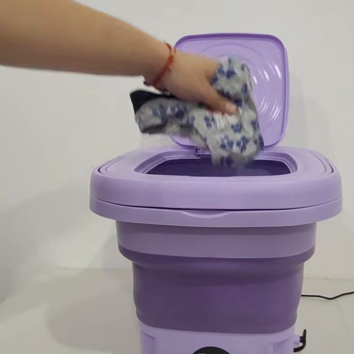 Una mano echa un par de prendas chicas a la mini lavadora lila armada, con la tapa abierta