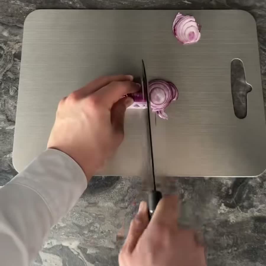 Una tabla de picar de acero inoxidable en uso sobre la mesada, vista desde arriba, con cebolla morada sobre su superficie